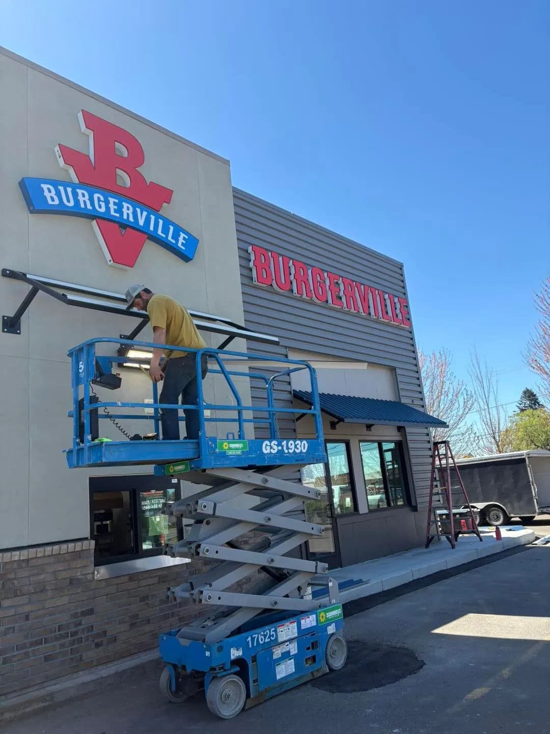 Worker installing sign for Burgerville using a lift outside the restaurant. Clear blue sky.