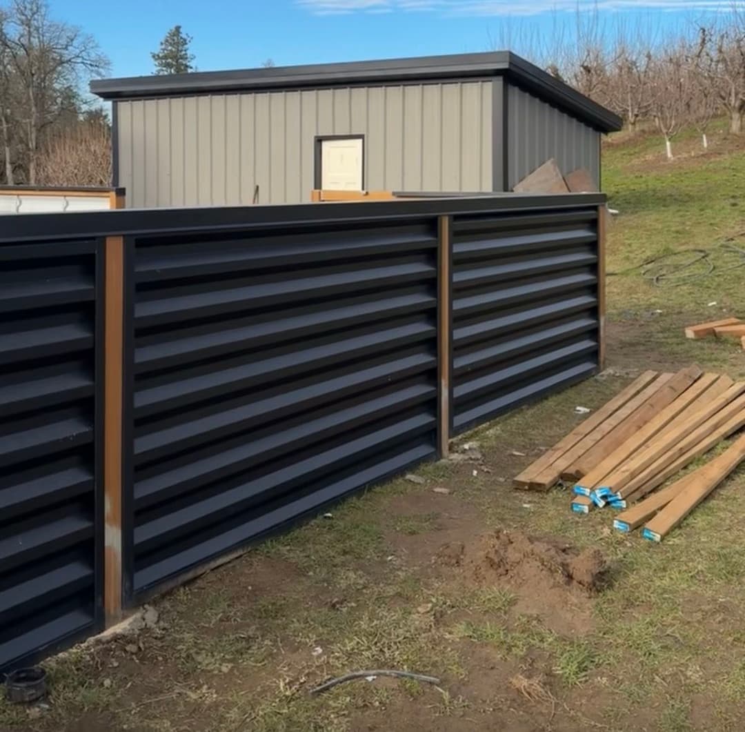 Modern black metal fence with wooden accents in a landscaped yard. Building materials nearby.