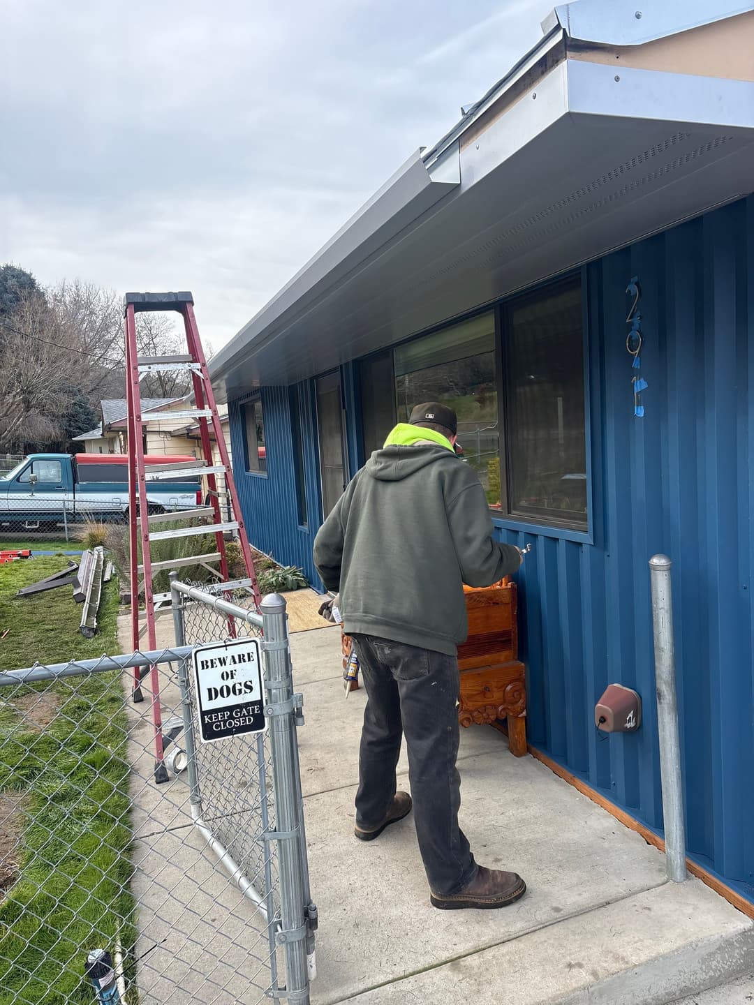 Person working on wooden furniture outside a blue house with a ladder and "Beware Dogs" sign.