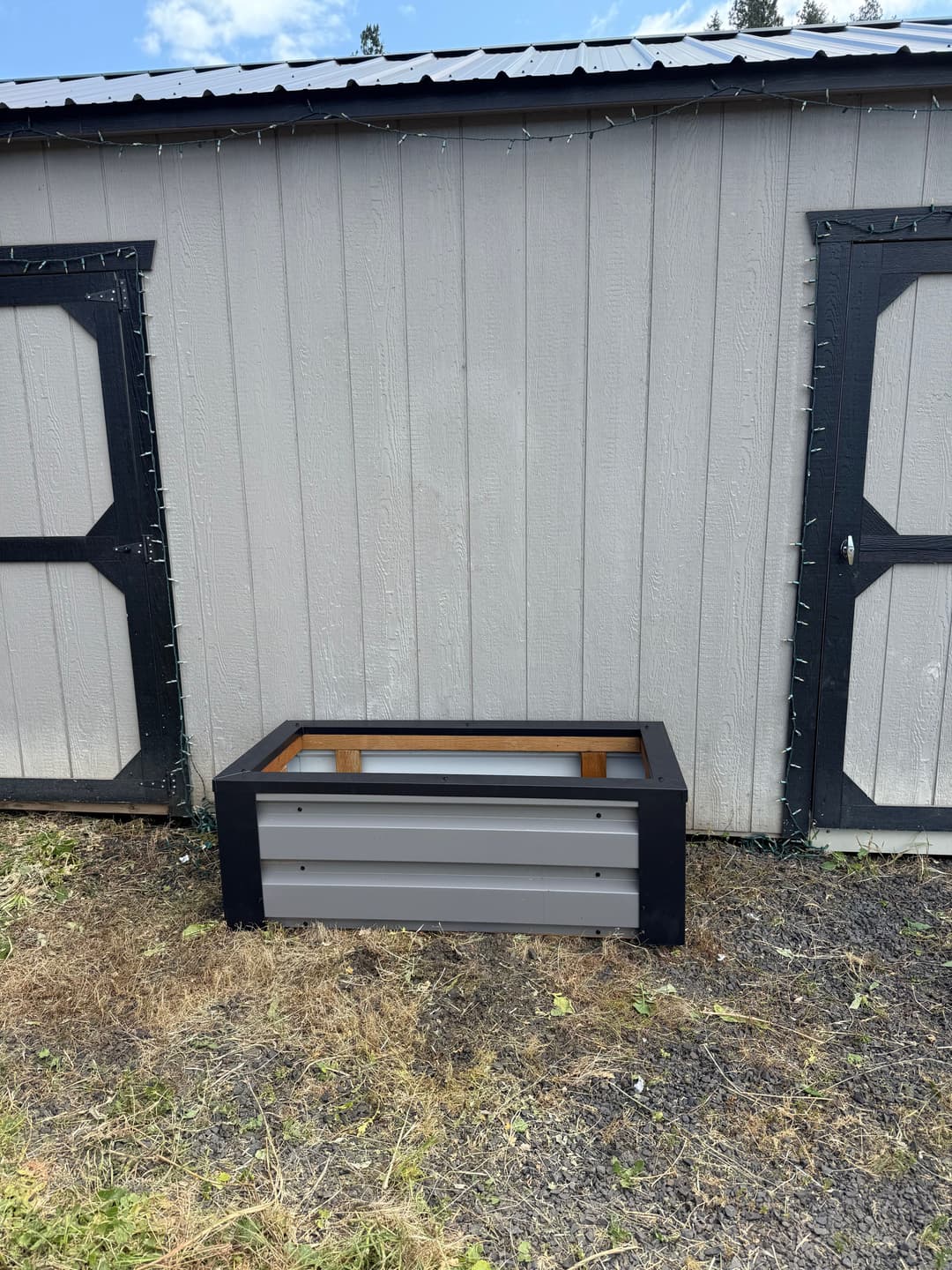 Outdoor raised garden bed in front of a shed with gray and black paneling. Ideal for planting.