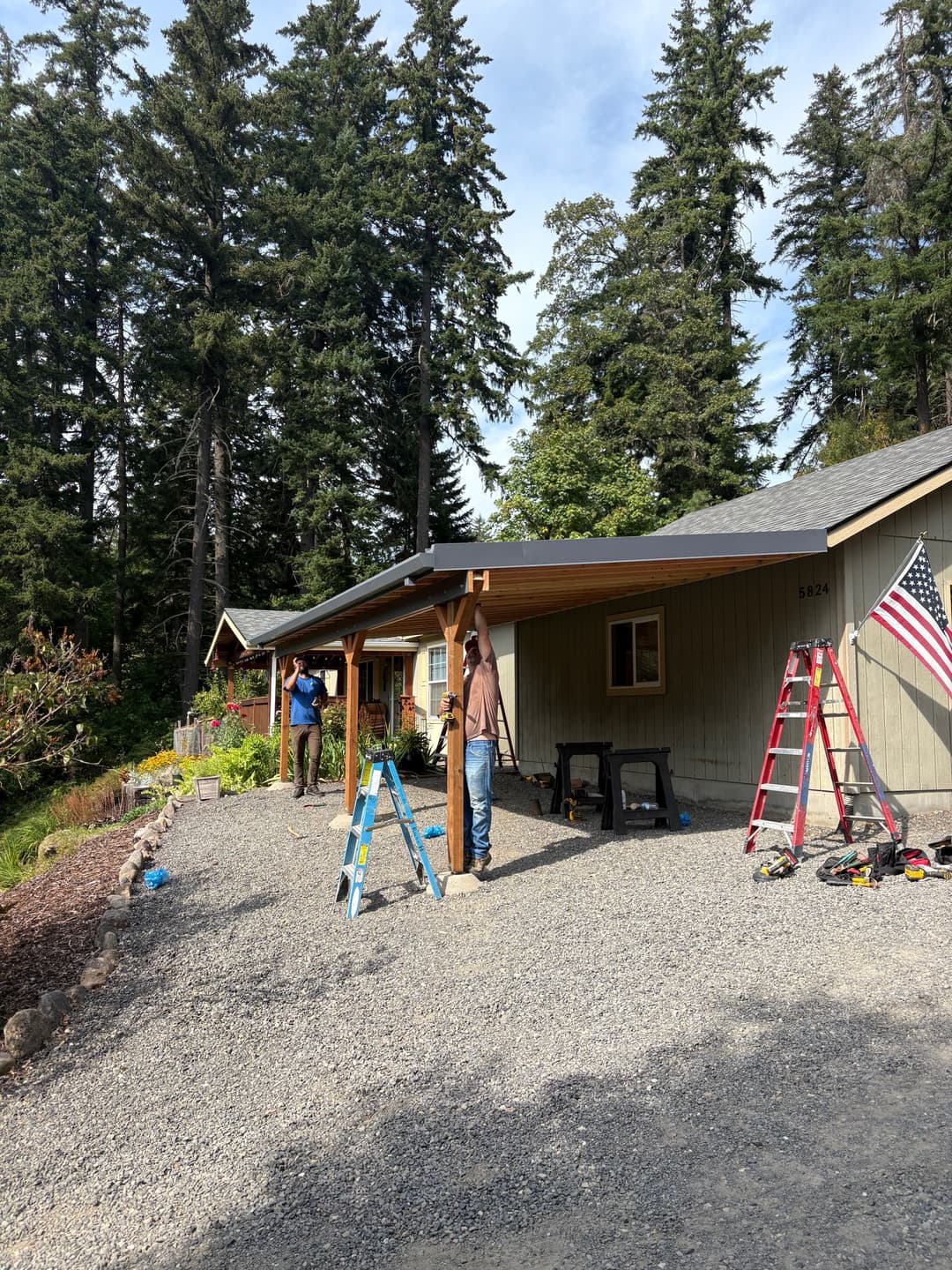 Workers installing an awning on a house surrounded by tall trees and tools.